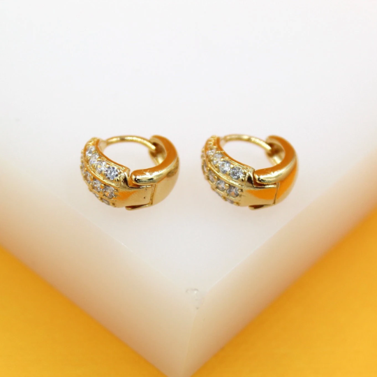 A pair of gold huggie hoop earrings embedded with sparkling cubic zirconia stones rests on a white display surface against a warm yellow background.
