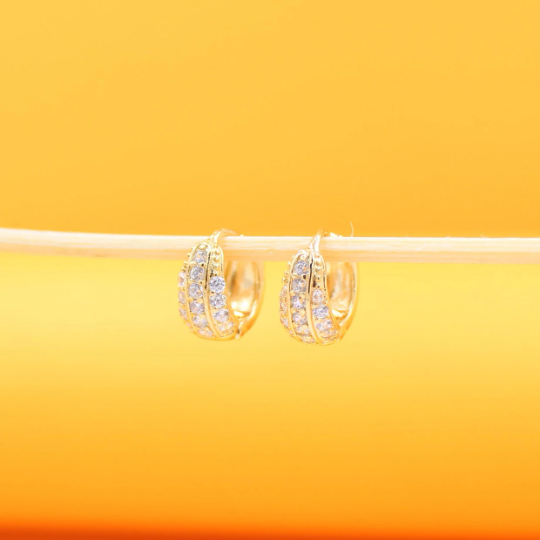Two sparkling gold pave hoop earrings rest delicately on a thin wooden stick against a warm yellow background.