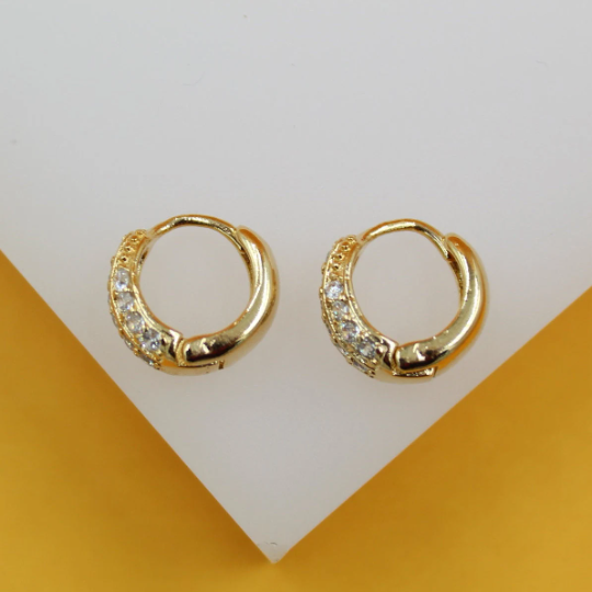 A pair of small gold huggie hoop earrings encrusted with sparkling cubic zirconia stones rests against a white display stand.