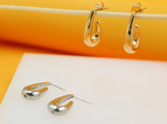 Two gold hoop earrings hang from a wooden stick above two silver hoop earrings resting on a white surface against an orange backdrop.