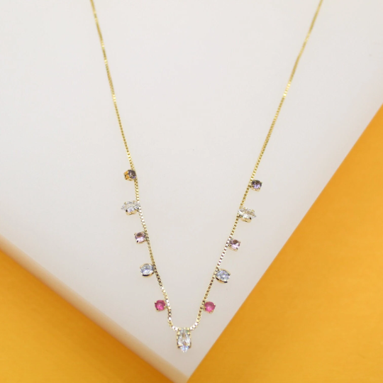 A delicate gold chain necklace dangles gracefully, featuring an array of colorful gemstones in various shapes and hues set along a white and orange display.