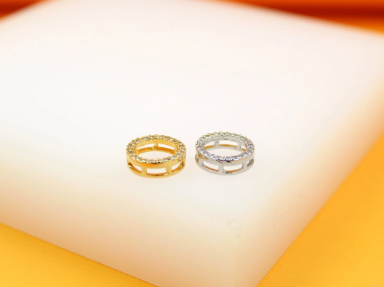 Two pave rhinestone round jewelry charms, one gold and one silver, sit side by side on a white display block.