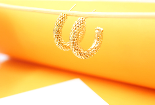 A pair of textured gold hoop earrings hangs delicately against a bright orange and white backdrop.