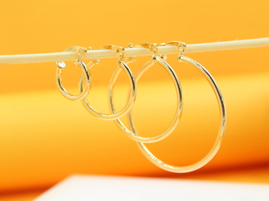 Four graduated gold hoop earrings hang neatly in a row from a thin wooden stick against a bright yellow background.