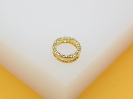 A small gold bail charm, encrusted with sparkling cubic zirconia crystals, rests on a white display block.