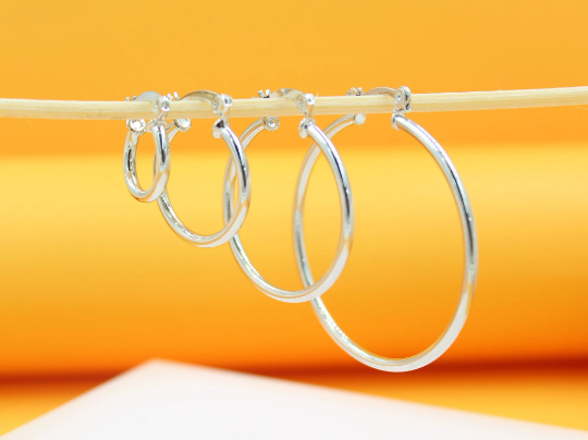 Four graduated silver hoop earrings hang in ascending size order on a thin wooden stick against an orange backdrop.