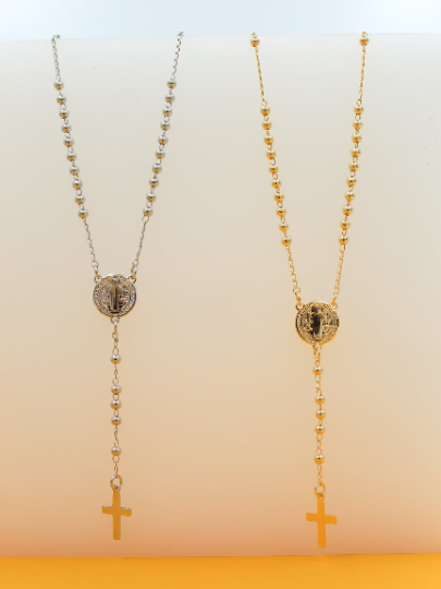 Two elegant Catholic rosary necklaces, one in silver and one in gold, hang side by side against a neutral white and orange backdrop.