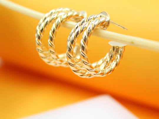 A pair of shiny twisted gold hoop earrings hangs delicately from a light-colored stick against a warm yellow and orange gradient background.