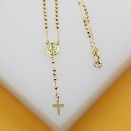 A delicate gold bead rosary necklace featuring a central Saint Benedict medal and a matching crucifix rests on a white display surface.