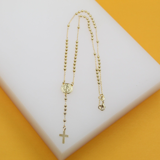 A delicate gold rosary necklace featuring bead chains, a central medallion, and a small cross rests on a white and yellow display surface.