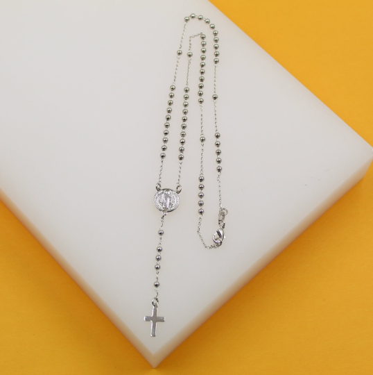 A delicate silver rosary necklace rests gracefully on a white display block against a vibrant yellow background.