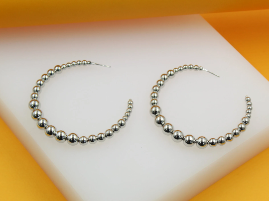 A pair of shiny silver beaded hoop earrings rests on a white and orange display.