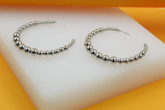 A pair of chunky silver beaded hoop earrings rests on a white and yellow display block.