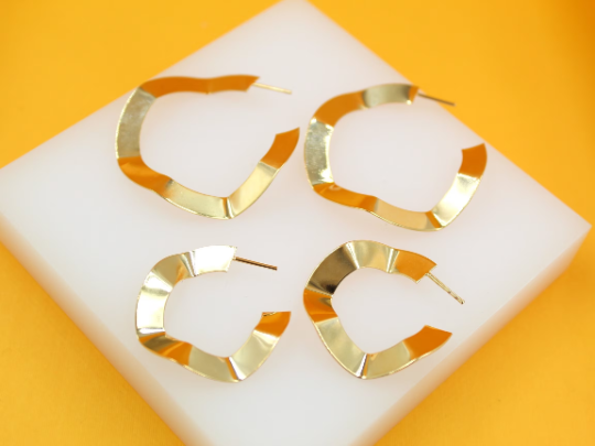Two pairs of wavy gold hoop earrings rest neatly on a white display square against a bright yellow background.