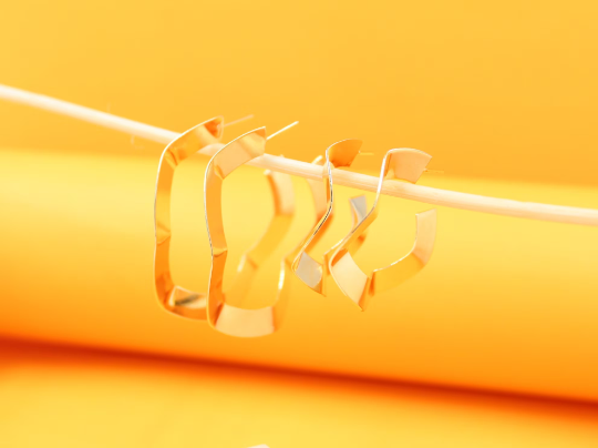 Two pairs of gold geometric hoop earrings hang delicately from a wooden stick against a bright yellow background.