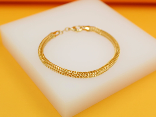 A delicate gold chain bracelet rests on a white display block against a vibrant yellow background.