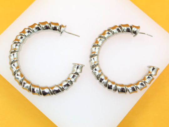 A pair of shiny silver hoop earrings featuring a unique twisted ribbon design rests on a white display against a vibrant yellow background.
