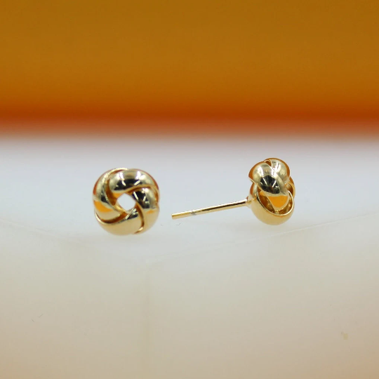 A pair of polished gold knot stud earrings rests on a white surface against an orange backdrop.