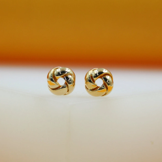 A pair of polished gold knot stud earrings rests on a smooth white surface against a warm orange background.