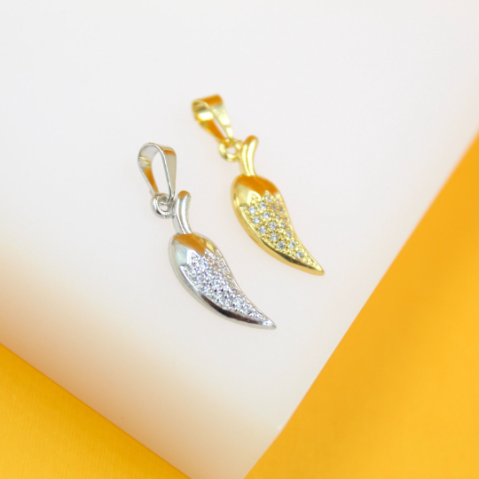 A silver and a gold chili pepper pendant, both encrusted with sparkling cubic zirconia stones, rest side-by-side on a white display surface against a vibrant orange background.