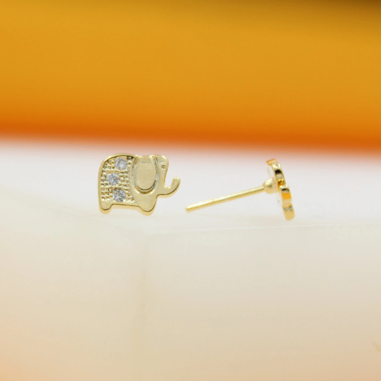 A pair of gold elephant stud earrings featuring sparkling crystal details rests against a soft white and orange background.