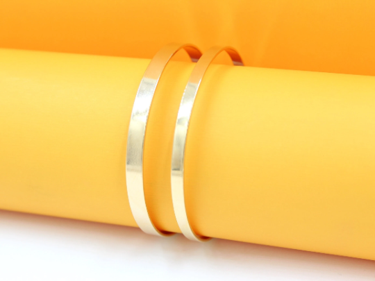 Two shiny gold bangle bracelets rest vertically on a bright yellow cylindrical display against a matching yellow backdrop.