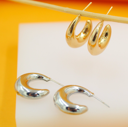 A pair of polished gold hoop earrings hangs delicately from a wooden stick above a pair of shiny silver hoop earrings resting on a white display surface.
