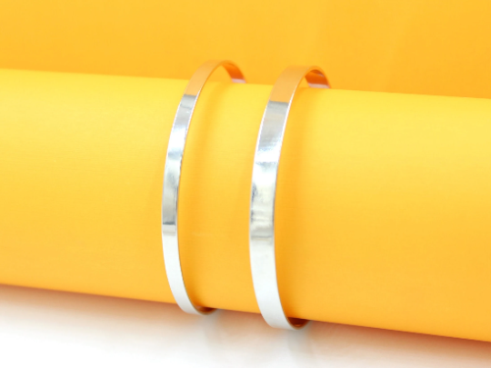 Two sleek, polished silver cuff bracelets rest on a vibrant yellow cylinder against a matching backdrop.