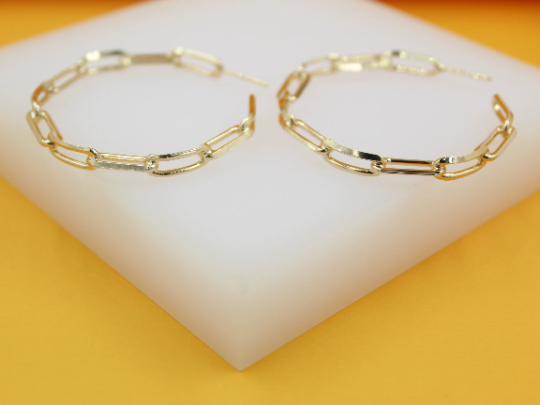 A pair of glossy gold paperclip link hoop earrings rests neatly on a white display stand against a vibrant yellow background.