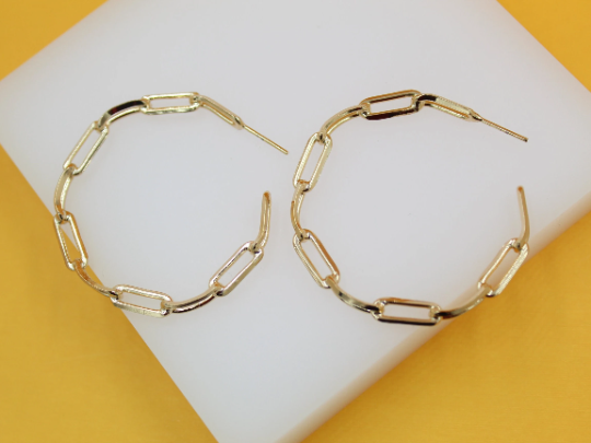 A pair of trendy gold paperclip chain hoop earrings rests on a white display surface.