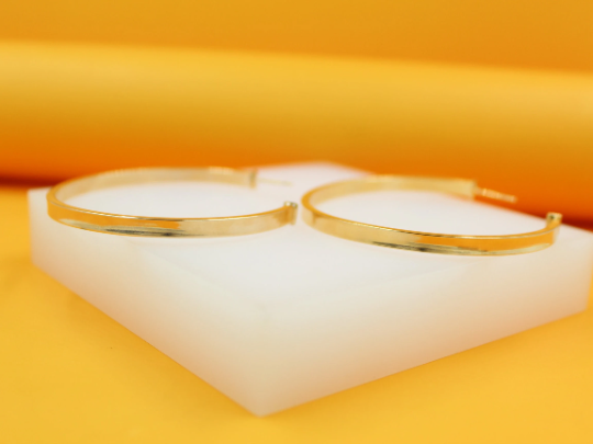 Two polished gold hoop earrings rest on a white display block against a vibrant yellow background.