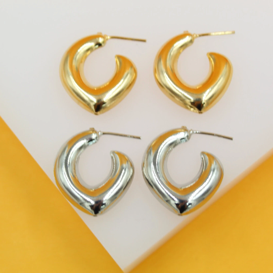 A pair of shiny gold heart-shaped hoop earrings rests above a matching pair of silver earrings on a white display surface.