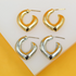A pair of shiny gold heart-shaped hoop earrings rests above a matching pair of silver earrings on a white display surface.