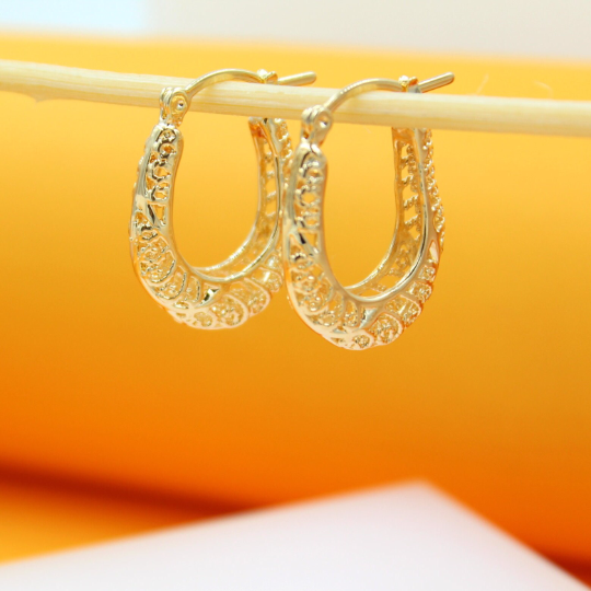 Two ornate gold hoop earrings hang delicately from a wooden stick against a bright orange and white background.