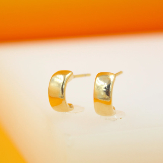 A pair of small, thick gold hoop earrings rest upright on a white display surface against a warm orange background.