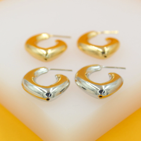 Two pairs of glossy gold and silver chunky heart-shaped hoop earrings sit neatly on a white display stand.