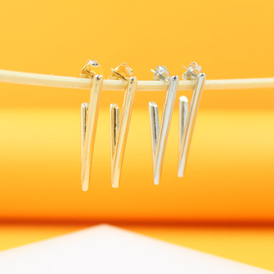 Four modern V-shaped hoop earrings in gold and silver hang delicately from a wooden stick against a bright yellow background.