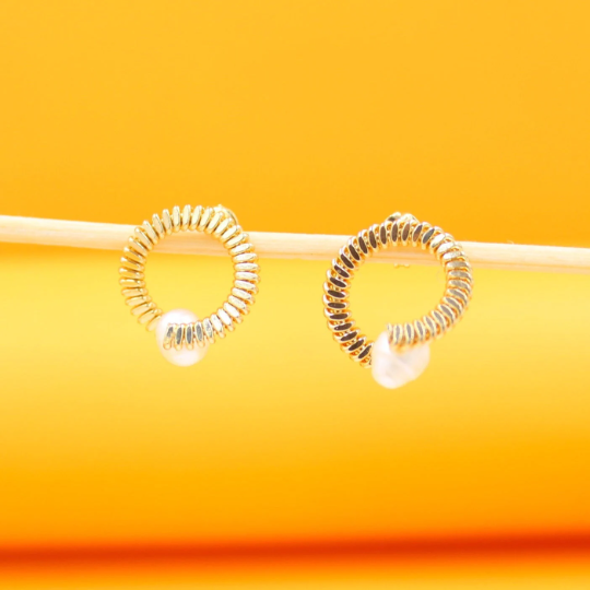 A pair of gold coiled hoop earrings featuring dangling pearls rest on a wooden stick against a bright yellow background.