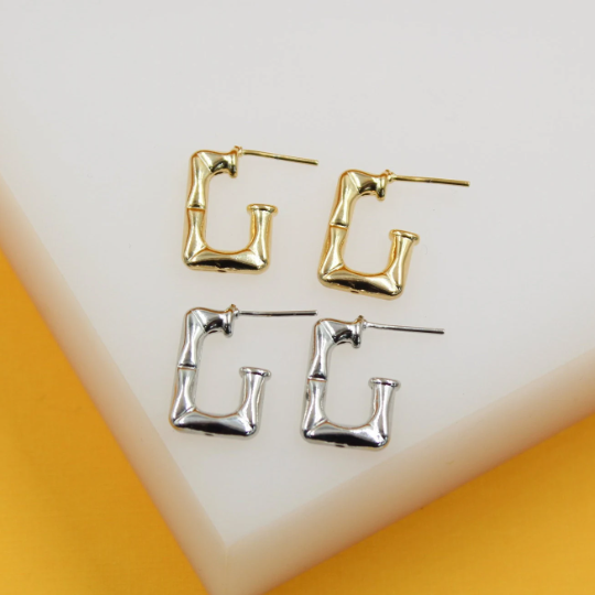 Two pairs of rectangular bamboo hoop earrings in gold and silver rest neatly on a white display stand.