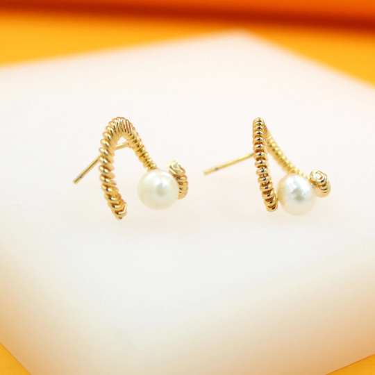 A pair of gold twisted rope earrings featuring dangling white pearls rests on a white display surface.