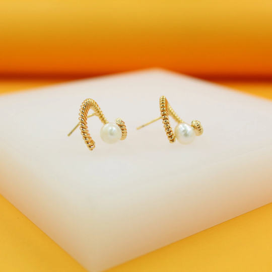 A pair of gold twisted rope earrings featuring a delicate white pearl rests on a white display block against an orange backdrop.
