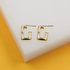 A pair of polished gold rectangular hoop earrings rests on a white display block against a vibrant yellow background.