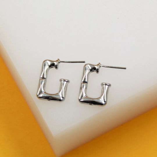A pair of silver rectangular hoop earrings with a textured bamboo design rests on a white display block.