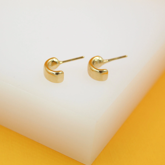 A pair of small gold huggie hoop earrings rests on a white and yellow display surface.
