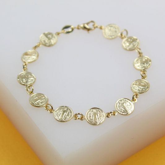 A delicate gold chain bracelet features linked Saint Benedict medals resting on a white display surface.