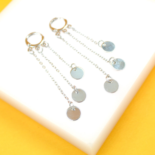 A pair of silver hoop earrings with cascading multi-strand chains and dangling coin charms rests on a white display surface against a vibrant yellow background.