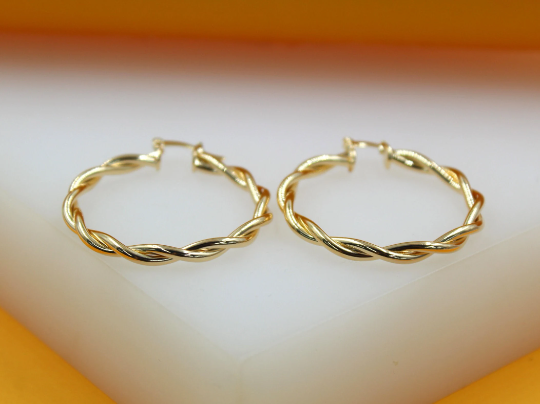 A pair of polished gold twist hoop earrings rests on a white display surface.