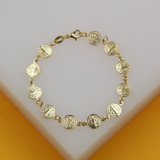 A delicate gold chain bracelet features linked Saint Benedict medals resting on a white display surface.