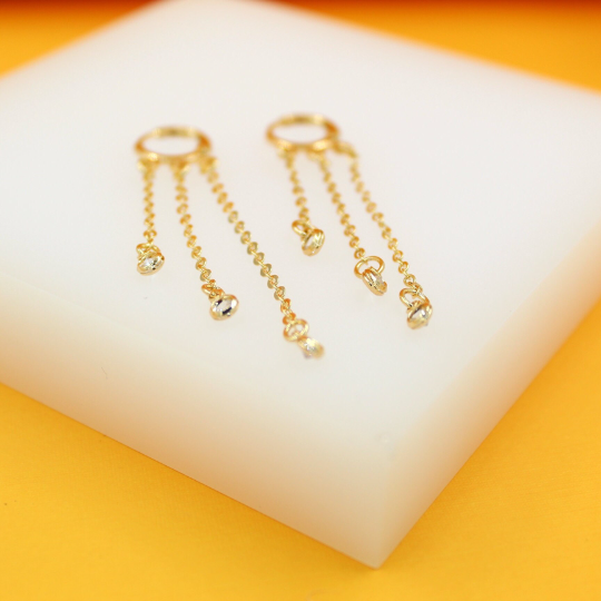 Two gold dangle earrings with triple hanging chains feature sparkling stone accents displayed on a white stand.
