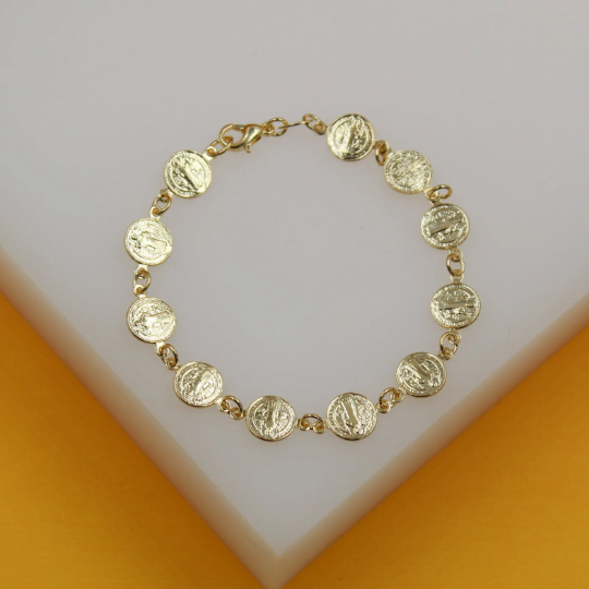 A delicate gold Saint Benedict coin charm bracelet rests on a white and yellow display surface.